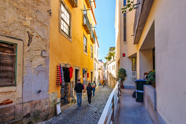 People walk down a narrow, cobblestone alley between colorful buildings with clothes displayed.