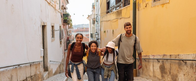 Four friends walking on a narrow cobblestone street between colorful buildings, smiling and arm-in-arm.