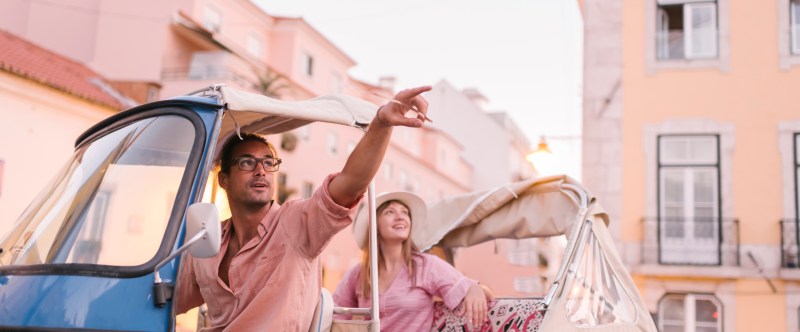 Two people in a small blue vehicle pointing and smiling in a European street.